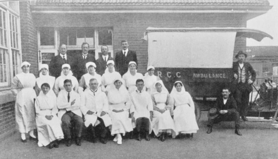[Image description: two rows of nurses in white gowns and a third row of men in black suits pose for a group photo in front of a vehicle marked “R.C.C. Ambulance”]Staff and ambulance outside Richmond State School emergency hospital